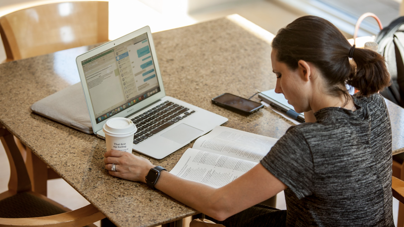 A woman sits at a table in front of a laptop and textbook