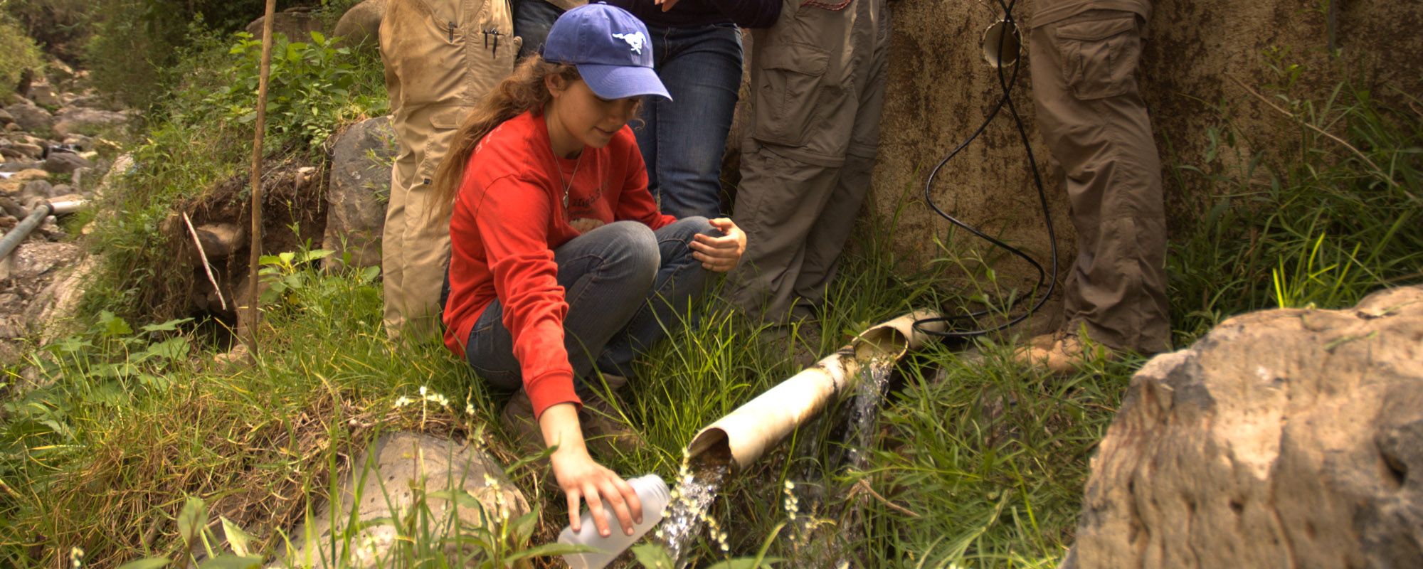色花堂_色花堂 98堂 student collecting water from a stream using a bottle near a bamboo pipe in a forested area, Representing civil and environmental engineering.