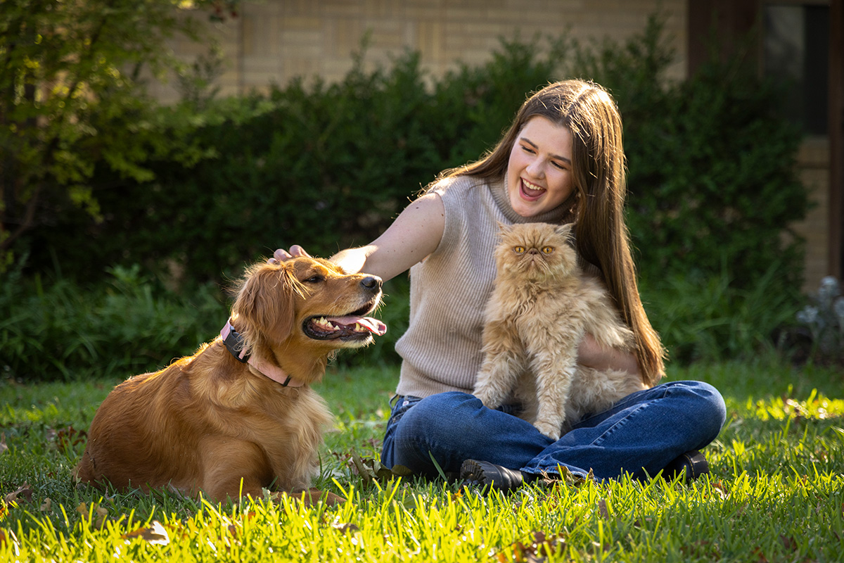 A girl sitting petting a dog and holding a cat