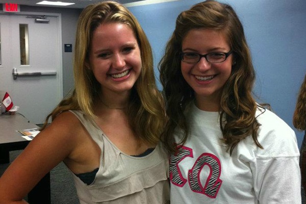 Two female students smile at the camera with an arm around each other. One student is wearing a t-shirt with sorority letters while the other student wears a light colored tank top.
