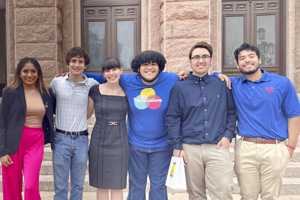 A group of six young adults stands together in front of a historic stone building with large windows and decorative trim. They are dressed in a mix of business casual and casual attire, including blazers, dresses, polo shirts, and jeans. They are smiling and standing closely with arms around each other, conveying a sense of camaraderie.