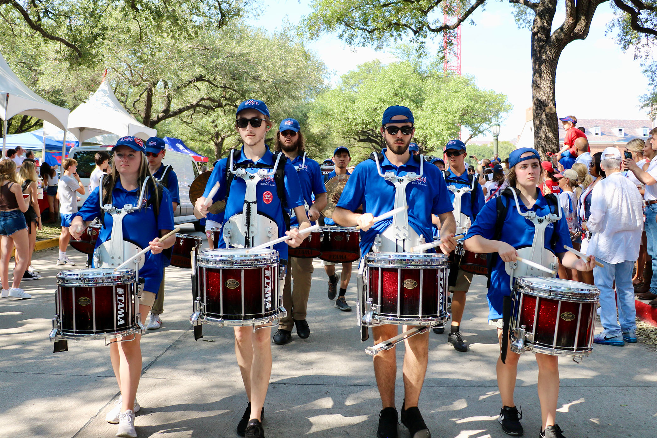 four band members in blue shirts playing snare drum marching outside down Bishop boulevard